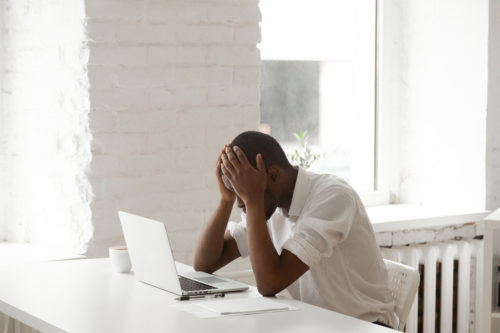 Overwhelmed man holding head at desk with laptop, stress from financial or legal issues.