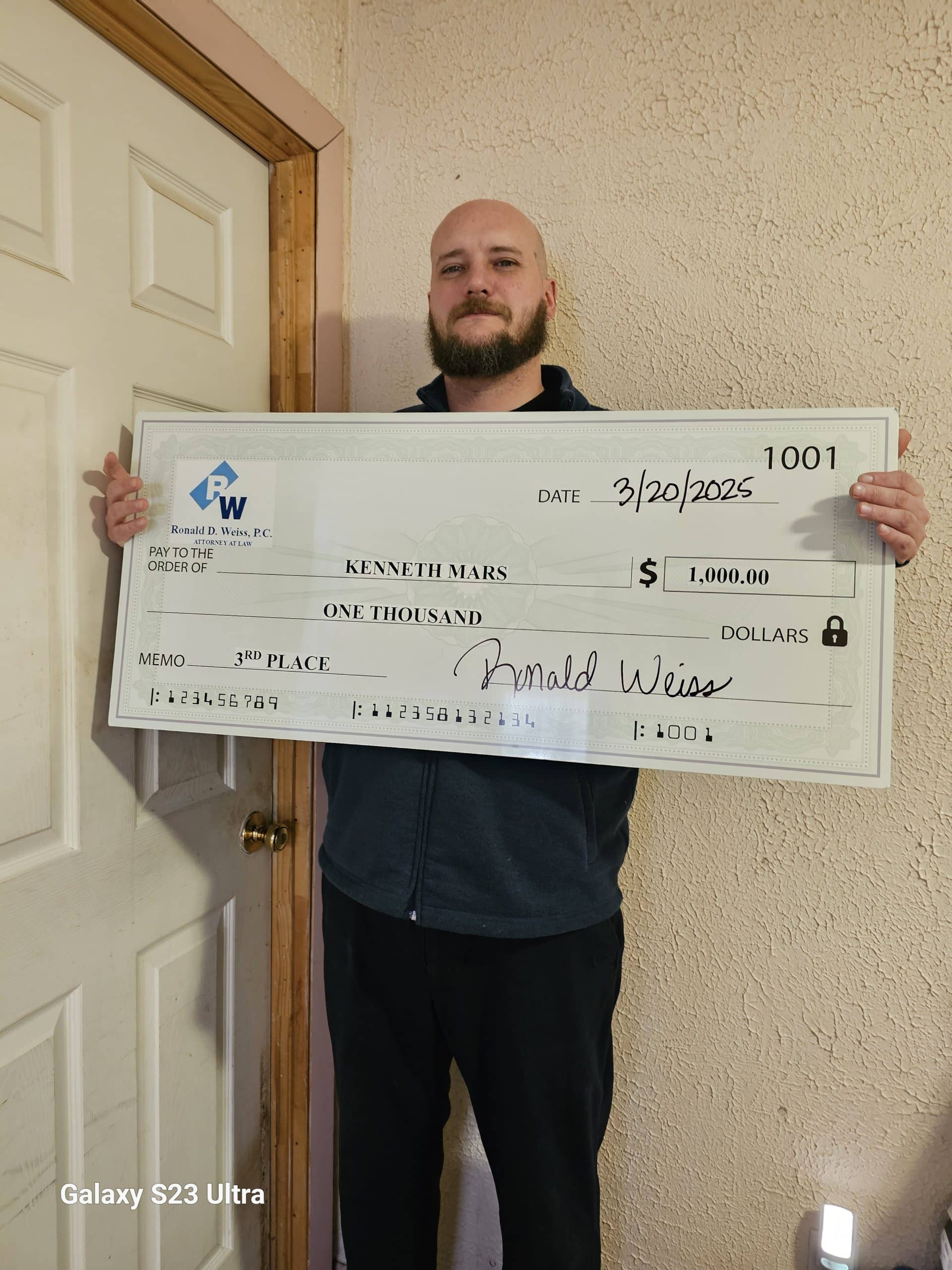 A man with a beard stands indoors, holding a large novelty check for $1,000 dated 3/20/2025, made out to Kenneth Mars for third place, signed by Ronald Weiss. The check is from Ronald D. Weiss, PC.