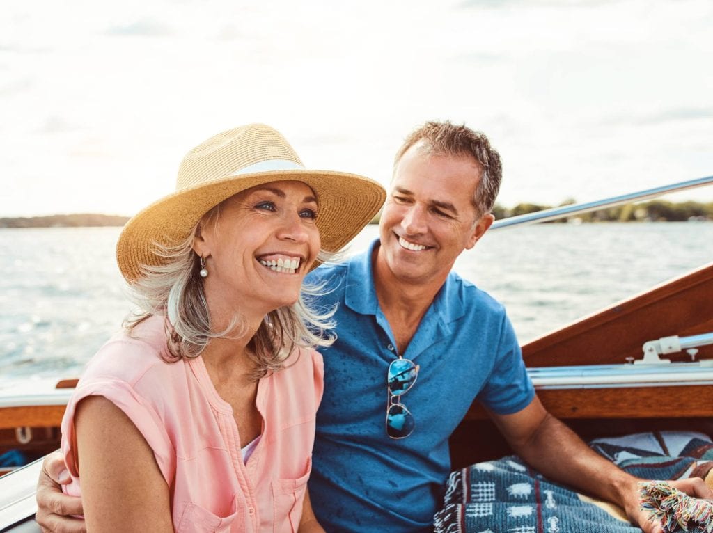A smiling older couple sits together on a boat, enjoying a sunny day on the water. The woman wears a straw hat and pink blouse; the man, in a blue shirt, looks at her warmly.