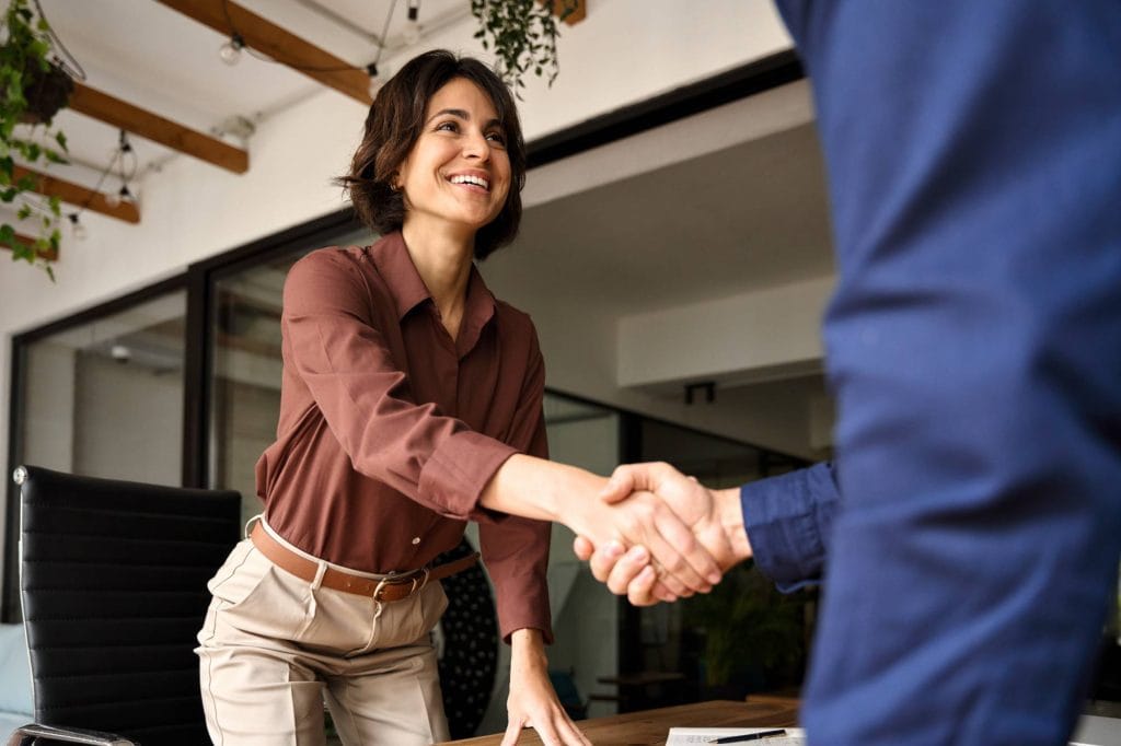 A woman in business attire smiles and shakes hands with another person across a desk in a modern office setting.
