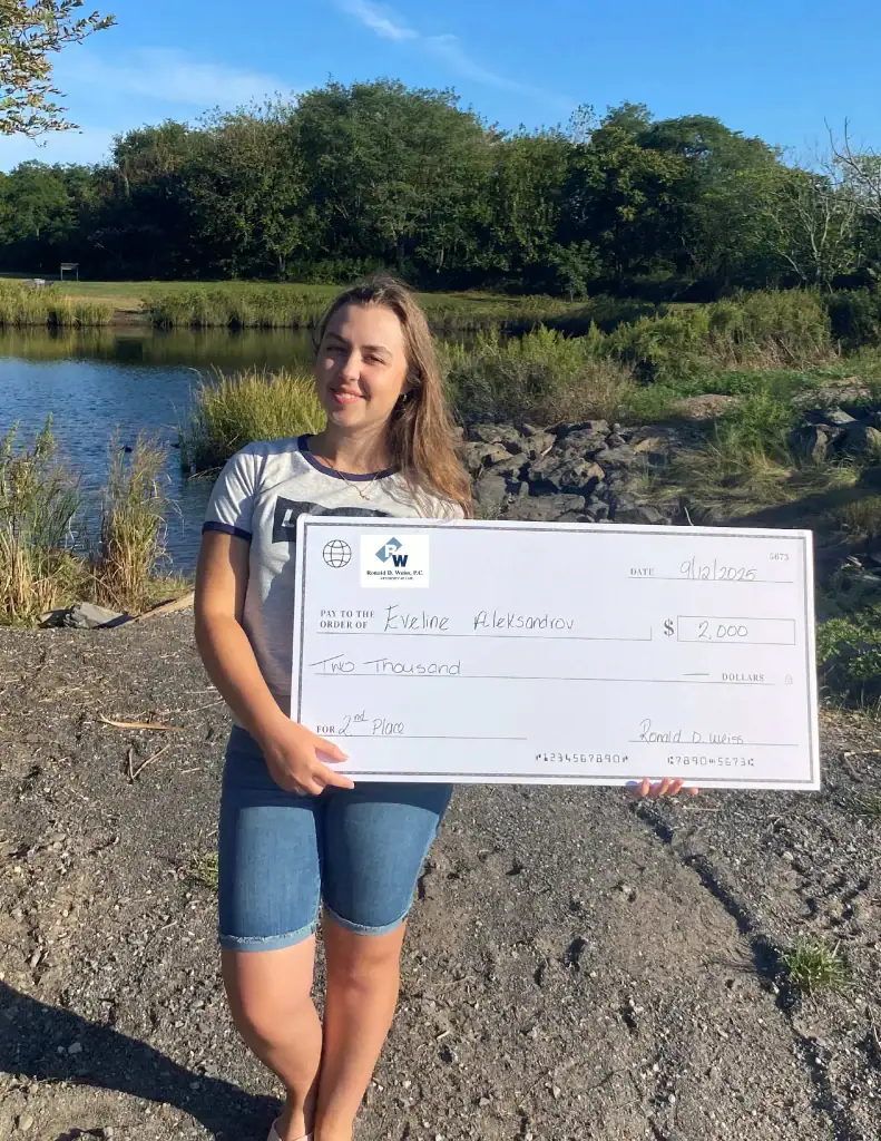 A young woman stands outdoors by a lake, holding a large check for $2,000 made out to Eveline Aleksandrov for 2nd place. She is wearing a casual t-shirt and shorts, smiling at the camera.