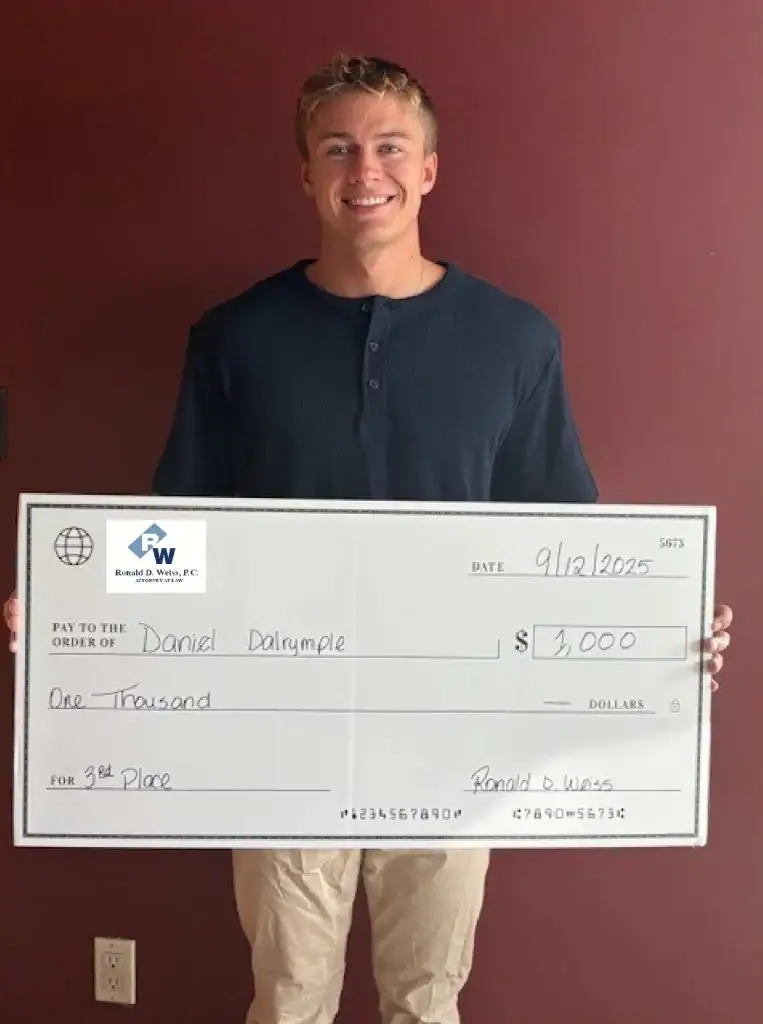 A smiling young man stands in front of a red wall, holding a large ceremonial check for $1,000 made out to Daniel Dalrymple for 3rd place, dated 9/12/2025 and signed by Ronald D. Weiss.