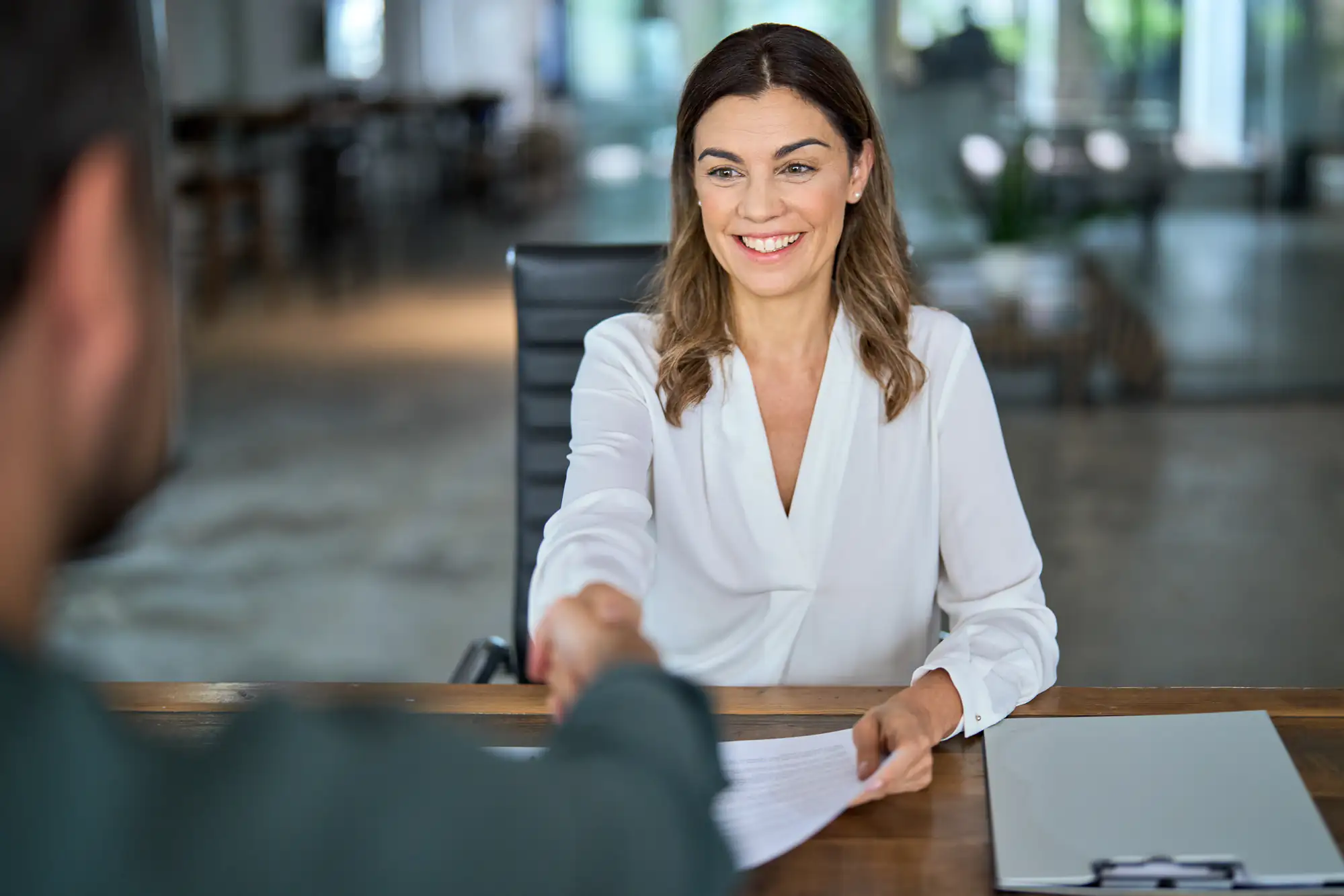 A woman in a white blouse smiles while shaking hands with someone across a desk in a modern office, holding a document. A closed laptop and papers are on the desk.