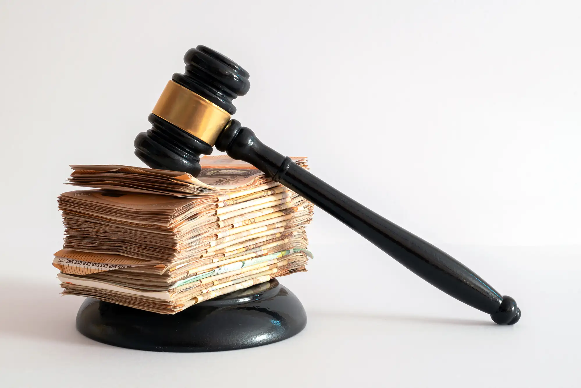 A judge's gavel rests on top of a large stack of paper currency, placed on a sound block, symbolizing the relationship between money and justice or legal decisions involving finances.