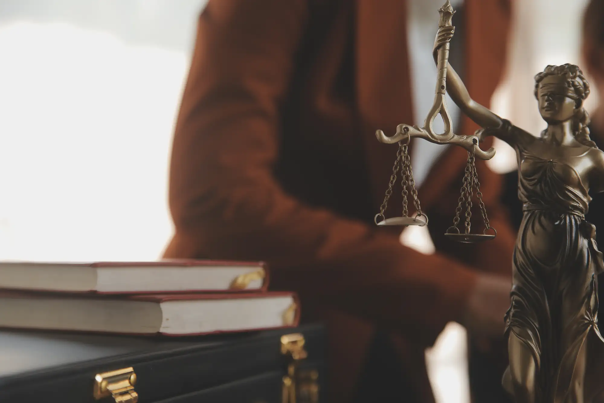 Close-up of a bronze Lady Justice statue holding scales, with two stacked books and a person in a brown blazer blurred in the background, suggesting a legal or law office setting.