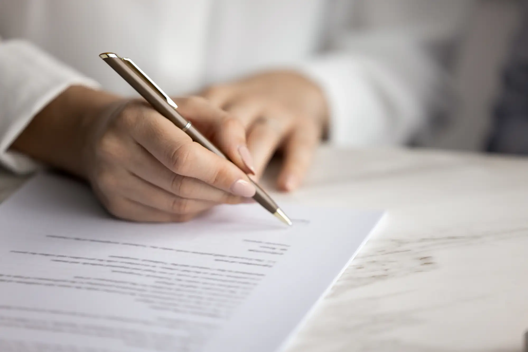 Close-up of a person&rsquo;s hand holding a pen and signing a document on a white marble table, with the rest of the document and background softly out of focus.