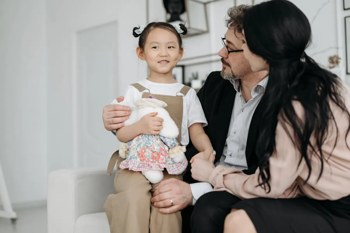 A young girl holding a stuffed toy smiles on a couch with two adults, enjoying family time indoors.