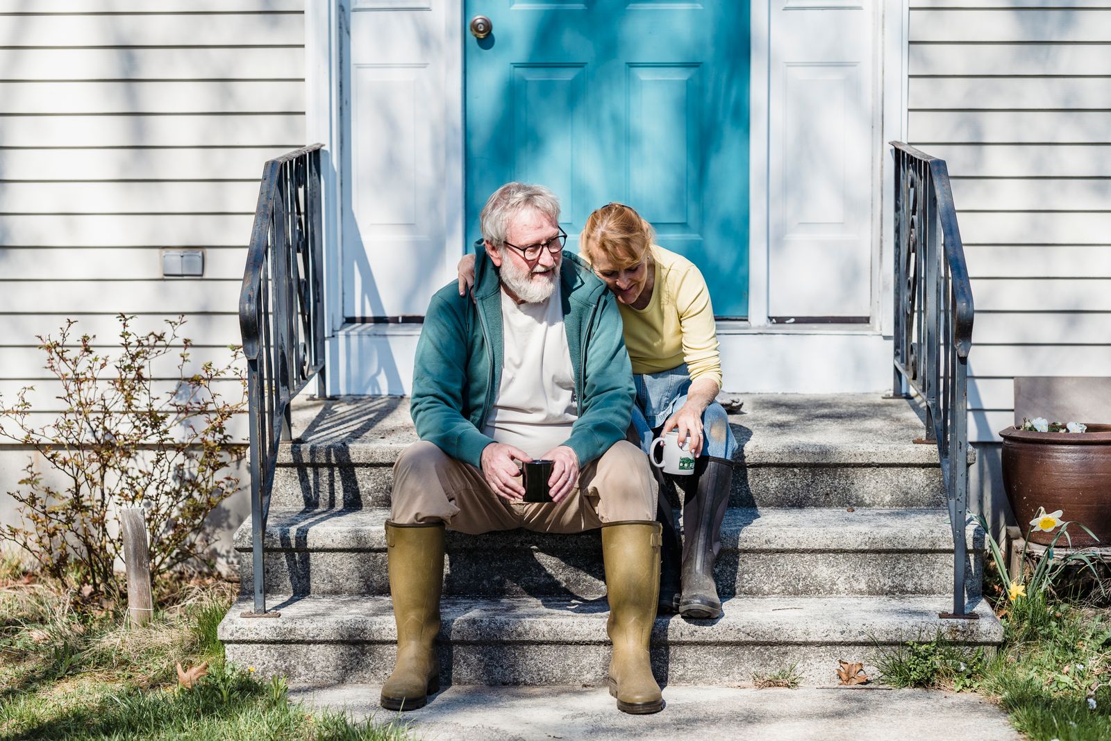 An older couple sitting affectionately on their home porch steps, sipping coffee together on a sunny day